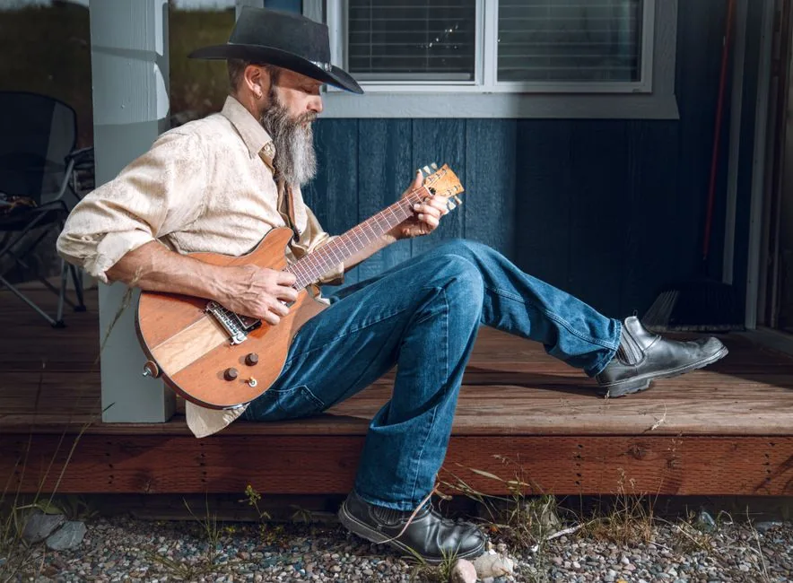 An elderly cowboy wearing a hat plays an electric guitar while sitting on a wooden porch.